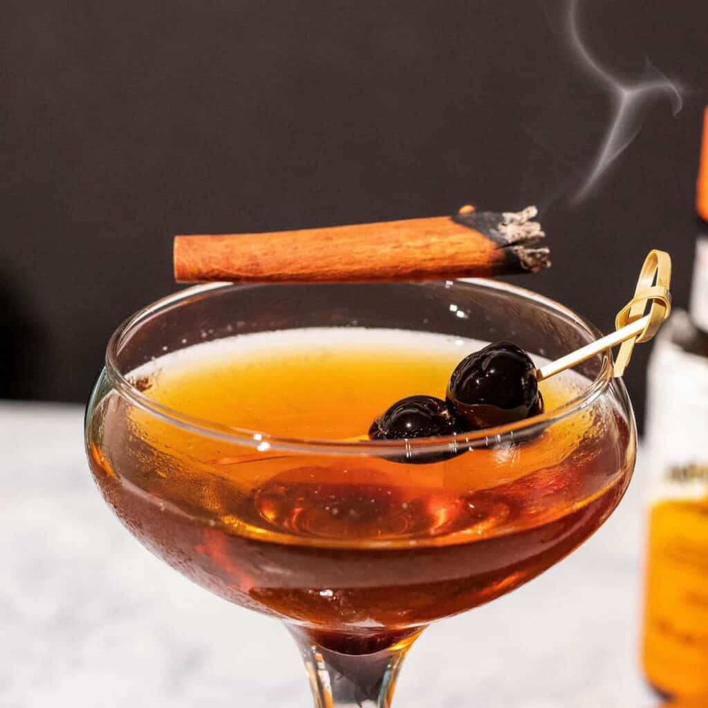 A cinnamon manhattan cocktail with a smoking stick of cinnamon on the rim 