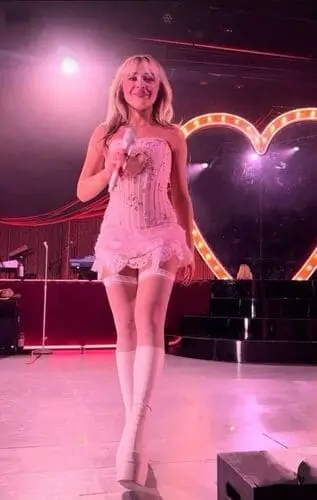 Sabrina Carpenter performing on stage in a stylish pink outfit, singing passionately into a microphone, with vibrant stage lights illuminating the background.