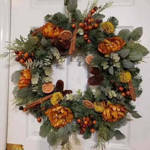 Fall wreath with eucalyptus and pine base, featuring dried orange slices, orange peonies, yellow allium, cinnamon bundles, and mixed greenery for a vibrant seasonal display.