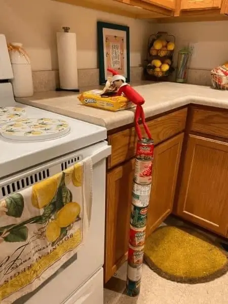 Elves on the Shelf building a tower of stacked cans to reach a package of Oreos, with festive Christmas decorations in the background.