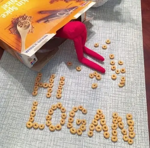 Elf on the Shelf digging into a Cheerios box, with the child’s name spelled out in Cheerios on the table, surrounded by festive holiday decor.