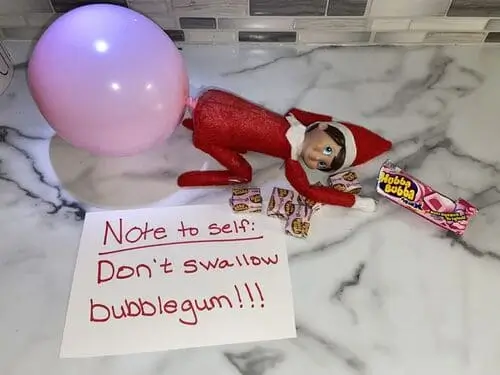 Elf on the Shelf surrounded by sticks of Hubba Bubba bubblegum with a note that says, "Note to self: Don’t swallow bubblegum," adding a humorous touch to holiday decor.