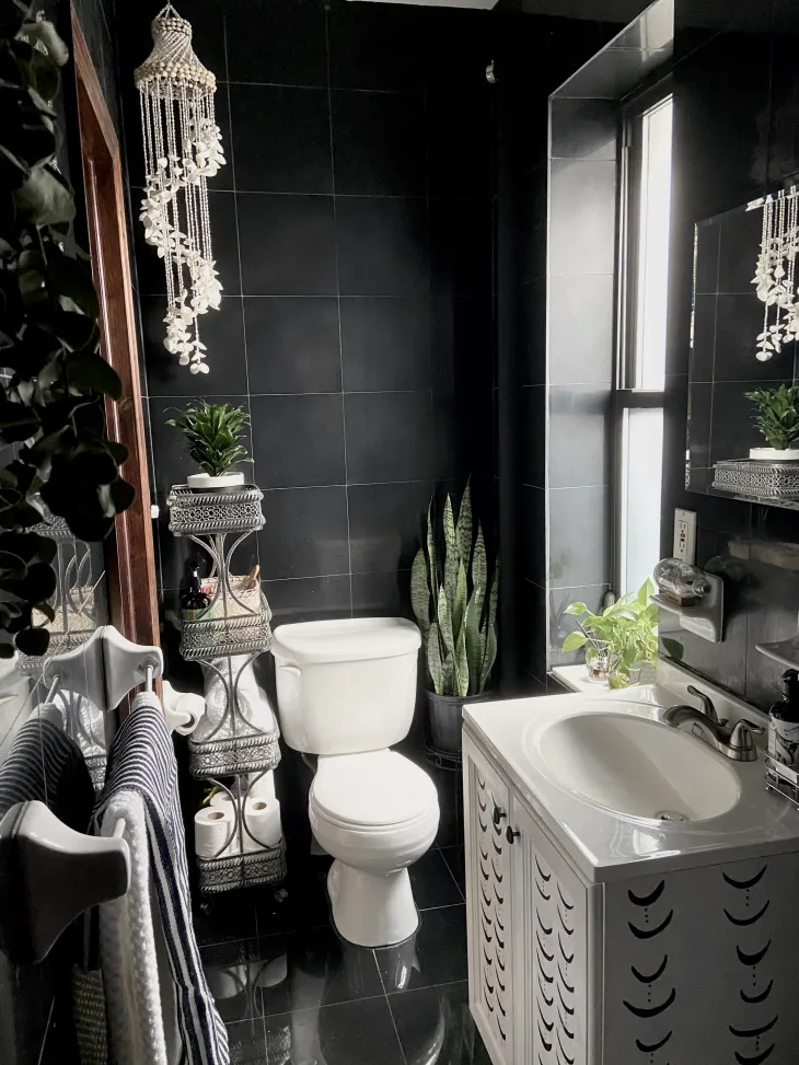 Dark moody small bathroom featuring white vanity with black crest design, moody tile walls, silver storage cart and towel racks, seashell hanging decor, and snake plants for an eclectic glam look.