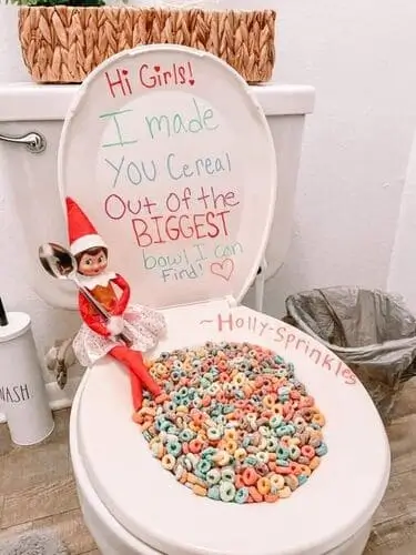 Elf on the Shelf holding a spoon with a toilet overflowing with Fruit Loops cereal and a dry erase note saying, "I made you a bowl of cereal out of the BIGGEST bowl I could find," surrounded by festive holiday decor.