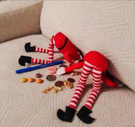 Elf on the Shelf bent over digging in couch cushions surrounded by loose change, candy wrappers, and a lost sock, with festive holiday decor nearby.