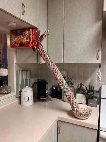 Elf on the Shelf sliding down a pool noodle "slide" coming out of a cereal box into a bowl of Fruit Loops, with the bowl secured in place using a sticky adhesive for a playful and colorful breakfast-themed holiday setup.