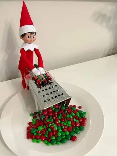 Elf on the Shelf grating colorful crayons into a bowl of Christmas M&Ms, surrounded by festive holiday decor.