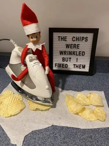 Elf on the Shelf ironing wavy potato chips into flat chips with a mini iron, next to a letterboard sign reading "The chips were crinkled but I fixed them," with festive holiday decor in the background.