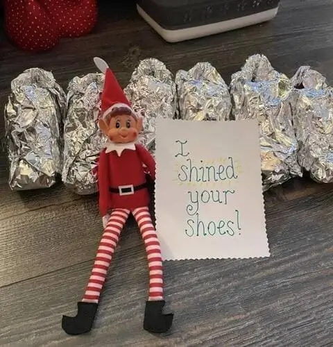 Elf on the Shelf next to kids' shoes wrapped in shiny tin foil with a playful note saying, "I shined your shoes!" and festive Christmas decorations in the background.