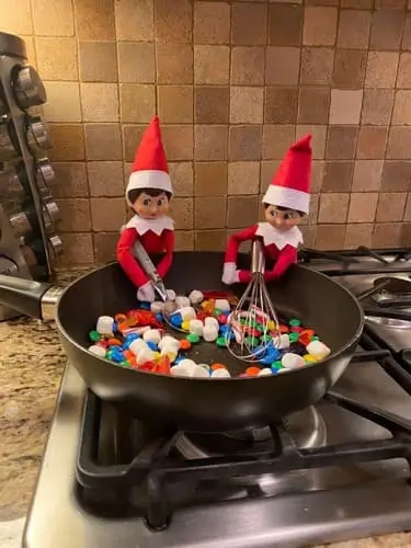 Elf on the Shelf making breakfast with M&Ms and mini marshmallows in a small bowl, surrounded by festive Christmas decorations.