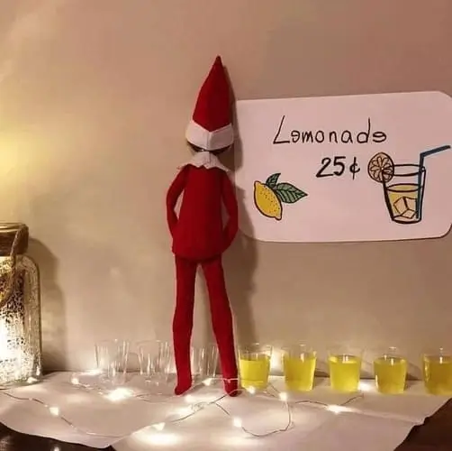 Elf on the Shelf leaning against a wall "peeing" lemonade into mini cups, with a handmade sign reading "Lemonade - 25¢," surrounded by festive holiday decorations.