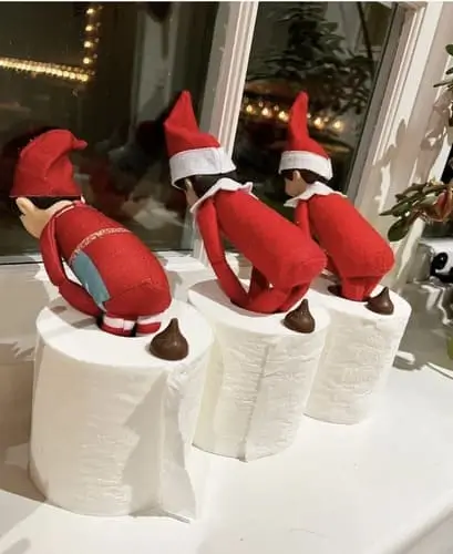 Elf on the Shelf in a roll of toilet paper, surrounded by Hershey Kisses, creating a funny holiday scene with a mischievous twist.