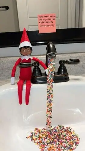 Elf on the Shelf by the sink with tape covered in sprinkles, creating the illusion of magic sprinkles flowing into the sink, surrounded by festive holiday decor.