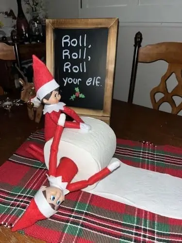 One Elf on the Shelf rolling another elf in a toilet paper roll with a chalkboard sign reading "Roll, roll, roll your elf... gently down the shelf," surrounded by festive holiday decor.