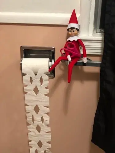 Elf on the Shelf balancing on a towel rack holding scissors, cutting toilet paper into snowflakes with festive holiday decor in the background.