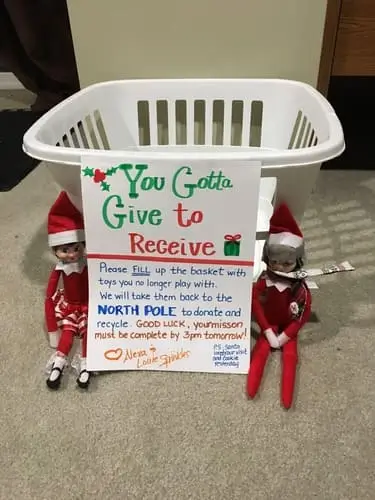 Elf on the Shelf near a laundry basket with a letter reading "You gotta give to receive," encouraging kids to donate old toys to the North Pole, surrounded by festive decorations.