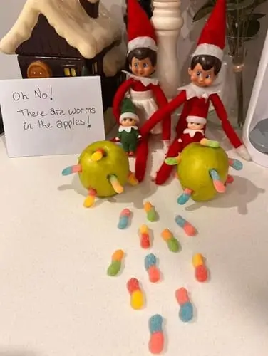 Elf on the Shelf beside apples with gummy worms crawling toward them and poking out of small holes in the apples, with a sign that says, "Oh no! There are worms in the apples!" placed on a kitchen counter for a playful holiday scene.