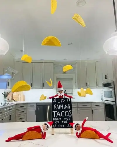 Two Elf on the Shelf dolls in taco shells, with one holding a chalkboard sign that reads "It's raining tacos from out of the sky," surrounded by taco shells hanging from the ceiling on fishing line to create a taco rain scene, surrounded by festive holiday decor.