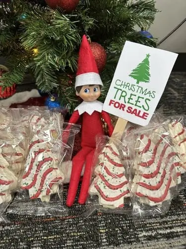 Elf on the Shelf next to a display of Little Debbie Christmas Tree Cakes with a "Tree for Sale" sign, surrounded by festive holiday decorations.