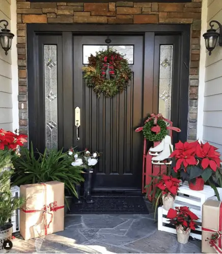 Farmhouse Christmas front door decorations with poinsettias, red sleigh, rustic wreath, kraft paper-wrapped gifts, rain boots with holly, and red plaid bow, perfect holiday entrance decor.