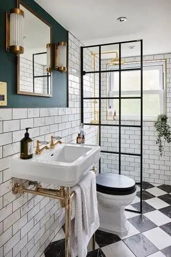 Victorian small bathroom with black-and-white checkerboard tile flooring, ornate toilet, black-framed shower, subway tiles, gold console sink, and elegant lighting.