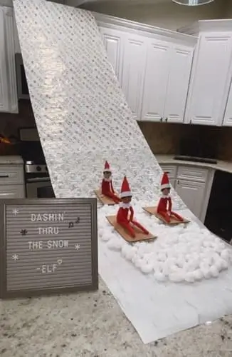 Elf on the Shelf sledding down a wrapping paper ramp from kitchen cupboards to the counter on cardboard sleds, landing in a cotton ball "snow" pile with a sign that says "Dashin' Through the Snow.