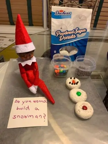 Elf on the Shelf next to powdered donuts stacked as a snowman with an orange M&M nose, mini chocolate chip eyes, and a note reading "Do you wanna build a snowman?" surrounded by holiday decor.