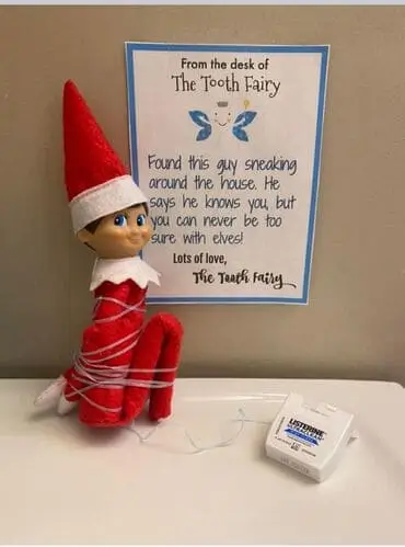 Elf on the Shelf wrapped in dental floss with a note that says, "Found this guy sneaking around the house. He says he knows you, but you can never be too sure with elves. Signed, The Tooth Fairy," surrounded by festive holiday decor.