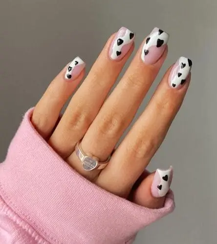 Valentine’s Cow Print Heart Nails with a sheer pink base, white swipes, and black hearts, inspired by the 2024 cow print trend popularized by celebs like Kendall Jenner.