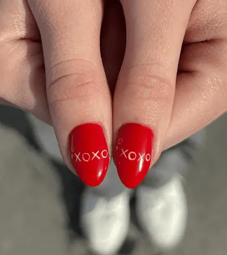 Valentine’s Day nail design with a mix of red and pink hearts on the pinkie, dainty heart with white dots on the index, swirly checkered print on the middle finger, cherries shaped like hearts on the ring, and bold red nails with 'XOXO' in pink on the thumbs for a fun and festive look.