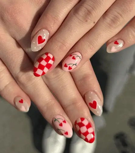 Creative Valentine’s nail design featuring red and pink hearts on the pinkie, dainty heart with white dotted accents on the index, swirly checkered print on the middle, cherries shaped like hearts on the ring, and red nails with 'XOXO' in pink on the thumbs for a playful and romantic look.